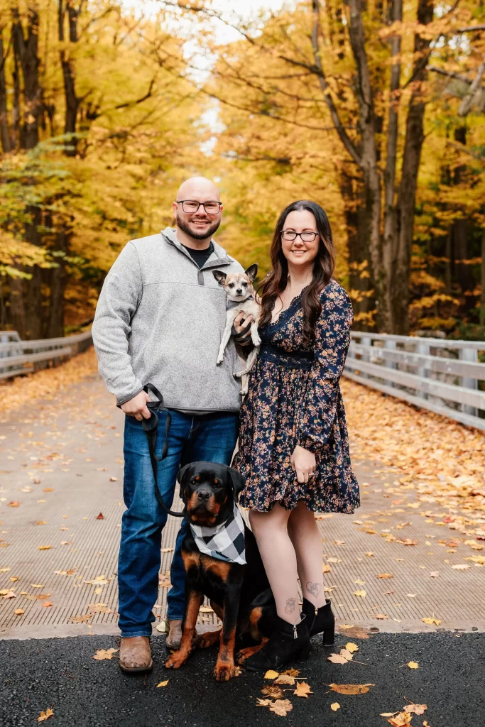 A couple with their two dogs on a bridge surrounded by autumn foliage.