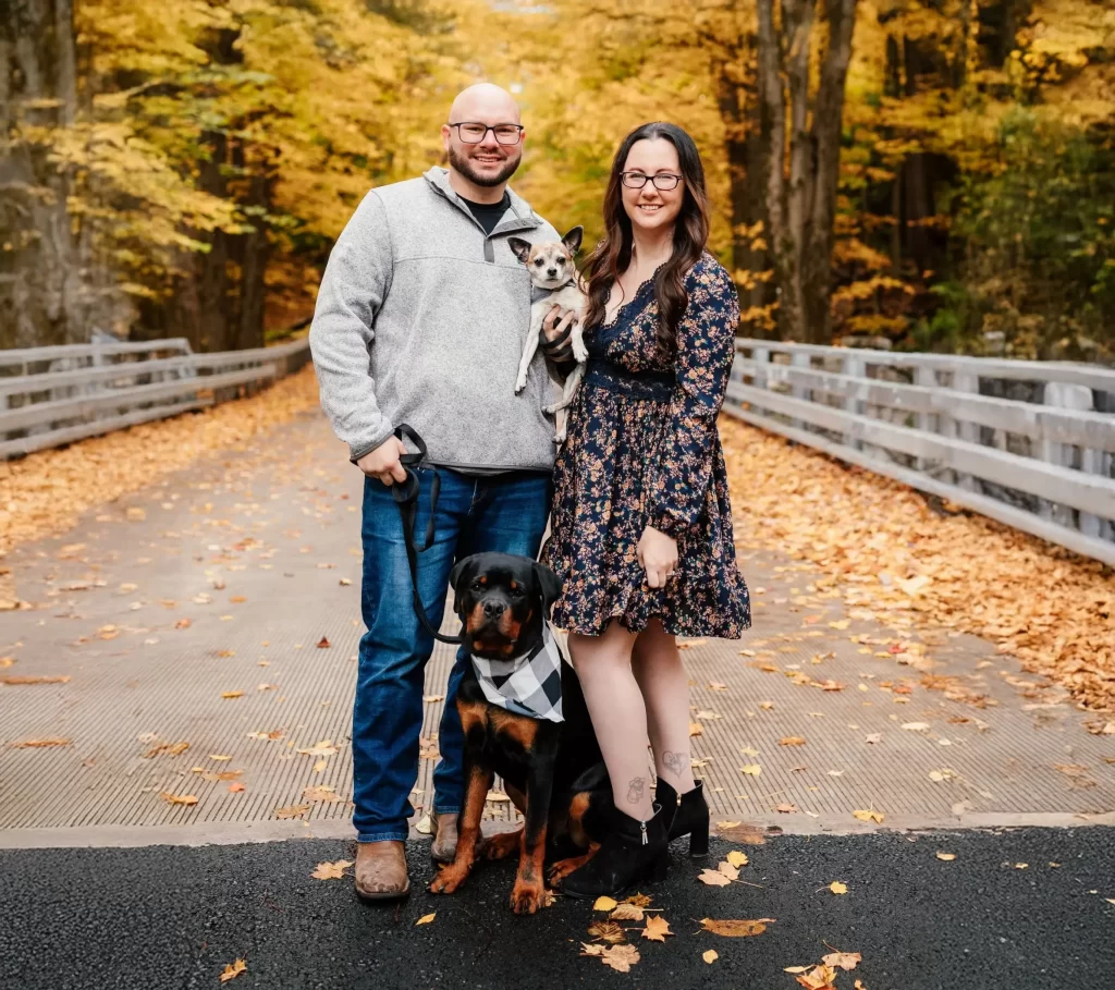 Couple standing with a small and large dog on a bridge surrounded by autumn leaves.