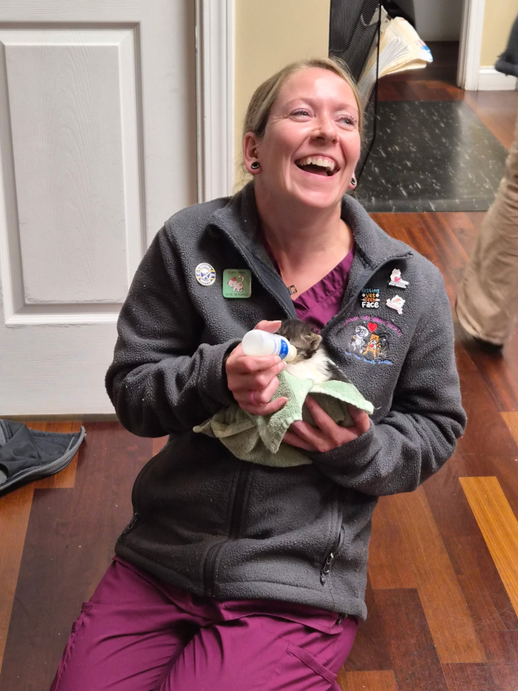 Woman laughing while holding and feeding a baby animal wrapped in a cloth.