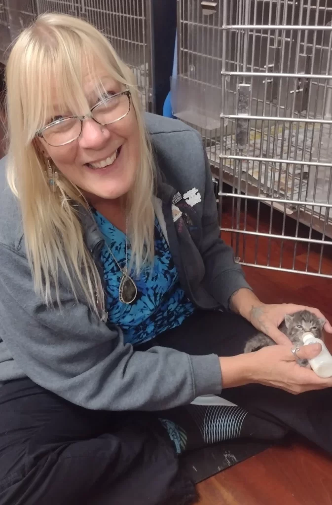 A smiling woman feeding a kitten with a bottle in a room with animal cages.